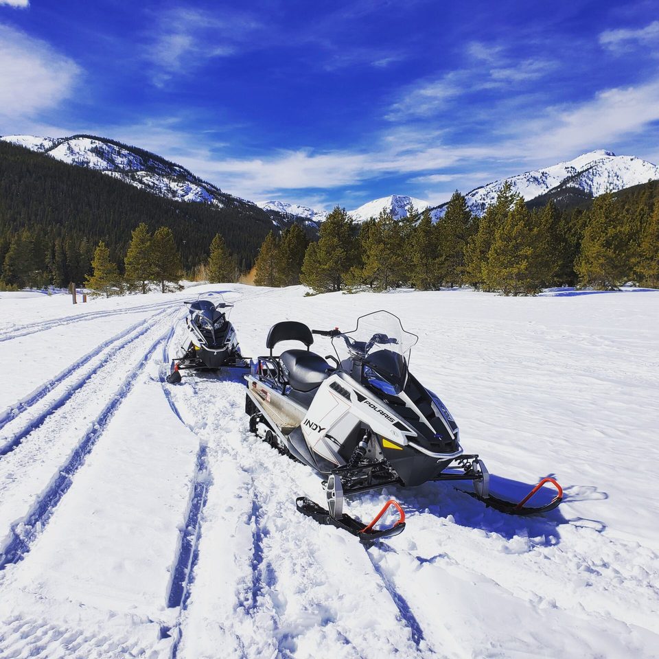 Two Polaris Indy snowmobiles parked on a groomed trail under a deep blue sky, snowy peaks behind the trees